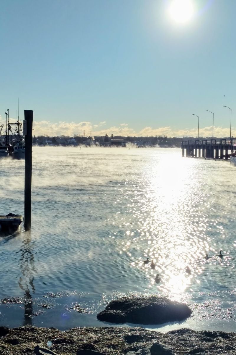 Early morning sun reflects on still water as fog lifts around boats at a working New England waterfront.